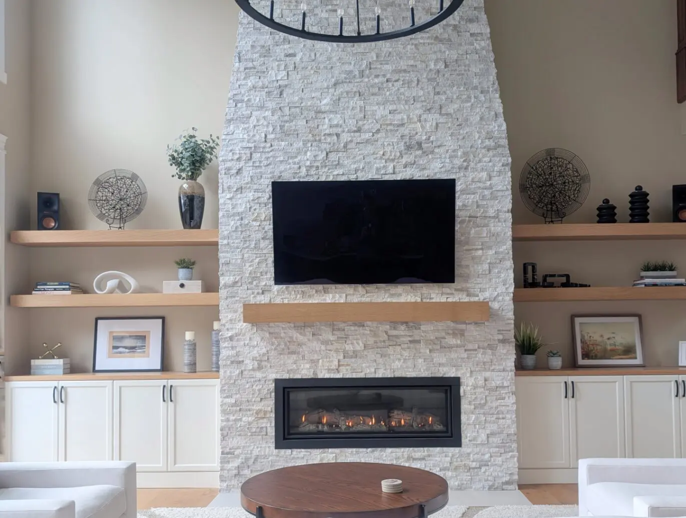 Modern living room by a transitional interior designer in Issaquah, WA, featuring a stone fireplace, wall-mounted TV, wooden shelves, cabinets, décor items, and a round coffee table.