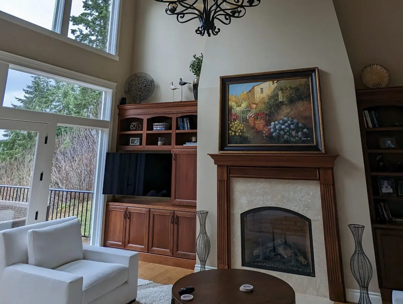 A living room designed by a transitional interior designer in Issaquah, WA, featuring a fireplace with a painting above the mantel, built-in shelves, a TV, white armchair, and large windows framing views of trees outside.