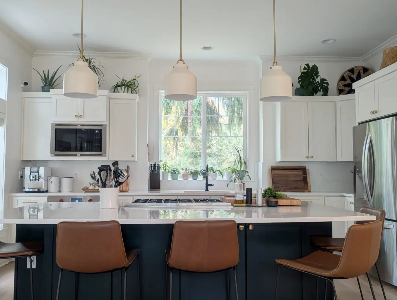 Modern kitchen with a large island, brown bar stools, white cabinets, stainless steel appliances, and pendant lights—styled by a transitional interior designer in Issaquah WA—with sunlight streaming through the window and lush plants.
