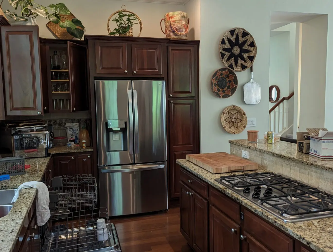 A modern kitchen by a transitional interior designer in Issaquah, WA, featuring dark wood cabinets, stainless steel refrigerator, gas stove, open dishwasher, and woven baskets on the wall. Boxes and kitchen items sit on the counter.