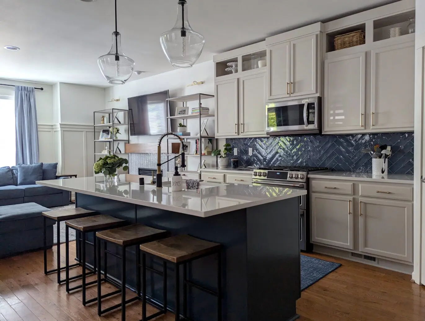 Modern kitchen by transitional interior designer Issaquah WA, featuring white cabinets, a dark island with four stools, stainless steel appliances, and a blue herringbone tile backsplash. Spacious living area with sectional sofa visible in the background.